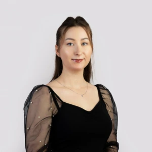 A young woman with long, straight brown hair, wearing a black blouse, with her arms lowered, smiling professionally, photographed against a neutral background.