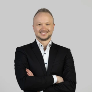 A young man with short dark hair, wearing a dark buttoned blazer and light shirt, arms crossed in front, smiling professionally, photographed against a neutral background.