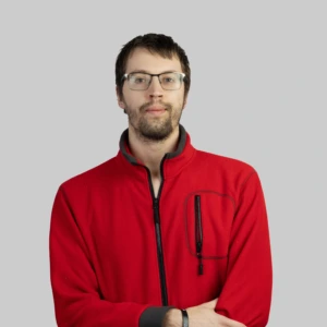 A young man with short dark hair, wearing glasses and a beard, dressed in a red zip-up fleece, standing with his arms crossed, calm and neutral expression, photographed against a neutral background.