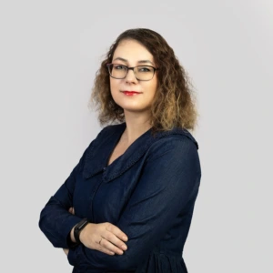 A young woman with shoulder-length brown hair, wearing a dark blue dress, arms crossed in front, smiling professionally, photographed against a neutral background.
