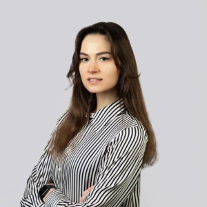 A young woman with long, straight brown hair, wearing striped shirts, arms crossed, smiling professionally, photographed against a neutral background.