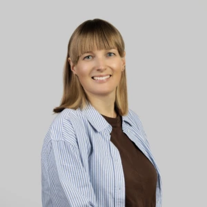 A young blonde woman with straight, shoulder-length hair, wearing blue striped button-up shirt, a brown blouse, smiling professionally against a neutral background.