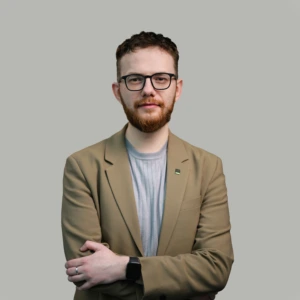 A young man with short dark hair, a short beard, wearing dark-framed glasses, a light brown blazer and gray shirt, arms crossed in front, with a professional expression, photographed against a neutral background.