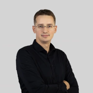 A young man with short, neatly styled dark hair, wearing glasses and a black shirt, standing with his arms crossed, calm and professional expression, photographed against a neutral background.