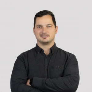 A young man with short dark hair, wearing dark shirt with the top button undone, arms crossed in front, with a professional expression, photographed against a neutral background.