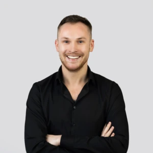 A young man with short dark hair, wearing black button-up shirt with the top buttons undone, smiling with a professional expression, photographed against a neutral background.