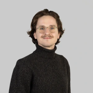 A young man with short dark hair, a mustache, and glasses, wearing a dark turtleneck sweater, smiling professionally, photographed against a neutral background.