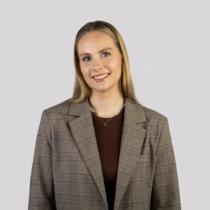 A young blonde woman with straight, shoulder-length hair, wearing a brown checkered blazer and a brown blouse, smiling professionally against a neutral background.