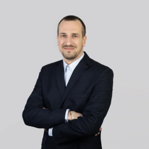 A young man with short dark hair, a mustache and beard, wearing a dark blazer and light shirt, arms crossed in front, smiling professionally, photographed against a neutral background.
