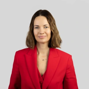 A young woman with shoulder-length brown hair, wearing a red blazer, smiling professionally, photographed against a neutral background.