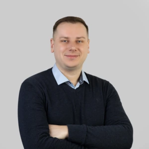 A young man with short dark hair, wearing a dark sweater and light shirt, arms crossed in front, smiling professionally, photographed against a neutral background.