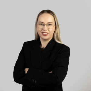A young blonde woman with straight, shoulder-length hair, wearing a black blazer, arms crossed, smiling professionally against a neutral background.