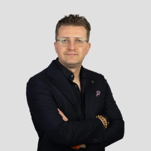 A young man with short dark hair, wearing glasses, a dark blazer and dark shirt, arms crossed in front, with a professional expression, photographed against a neutral background.