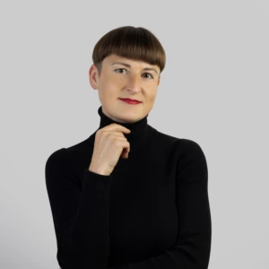 A young woman with short brown hair, wearing a black high-neck blouse, one hand raised near her chin and the other lowered, smiling professionally against a neutral background.