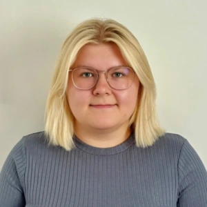 Portrait photograph – Jurgita Čėstauskaitė. A young woman with shoulder-length light blonde hair. She wears round glasses with transparent frames and a grey ribbed sweater. She looks directly at the camera with a calm, neutral expression. The background is light and neutral.