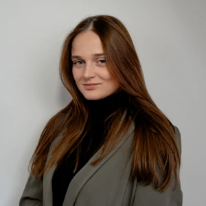 Roberta Kybartaitė Portrait photograph – Roberta Kybartaitė. A young woman with long straight brown hair. She is wearing a black turtleneck sweater and a grey-green blazer. She is slightly turned to the side while looking at the camera with a subtle smile. The background is light and neutral, studio-style.